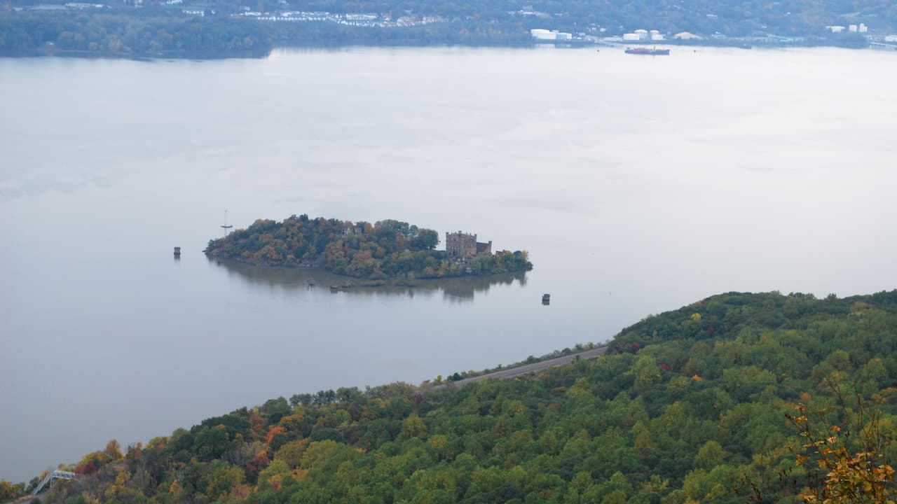 Looking down from Breakneck Ridge onto Pollepel Island in autumn: the ruined brick towers of Bannerman Castle stand among turning trees on the small island, old dock pilings in the water around it, a cargo ship anchored upriver and the far shore grey with haze.