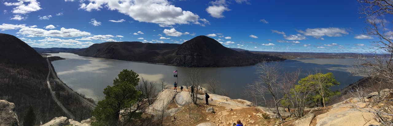 Panorama from the bare rock of Breakneck Lookout. The Hudson bends around a steep wooded headland far below; a few walkers stand at the edge of the outcrop on the right, giving the drop its scale.