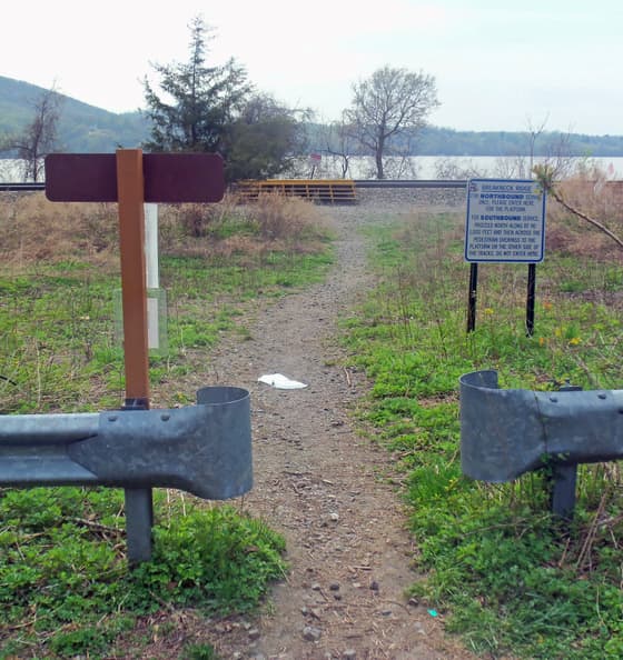 The footpath from the northbound Breakneck Ridge platform to the park: a rough stone-and-dirt track running between two lengths of steel highway guardrail, with a signpost and a notice board either side, and the river visible ahead through bare trees.