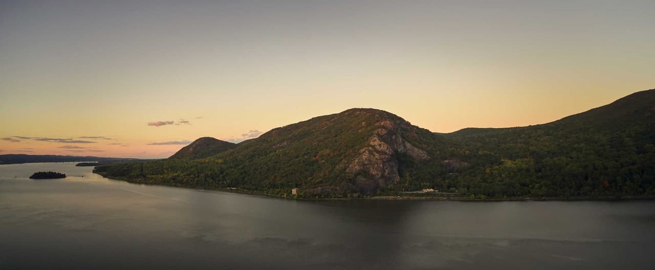 Wide panorama of Breakneck Ridge at sunrise from across the river: first light picks out the bare rock face above the water, the rail line threads along the shore at its foot, Pollepel Island sits small at the left edge, and the sky fades from peach to grey.