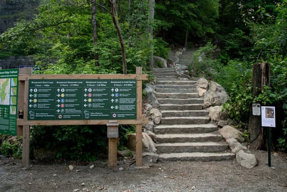 The Breakneck Ridge trailhead: a wide green information board carrying four route panels with times and distances, mounted on timber posts beside a flight of rough stone steps that climbs into the trees.