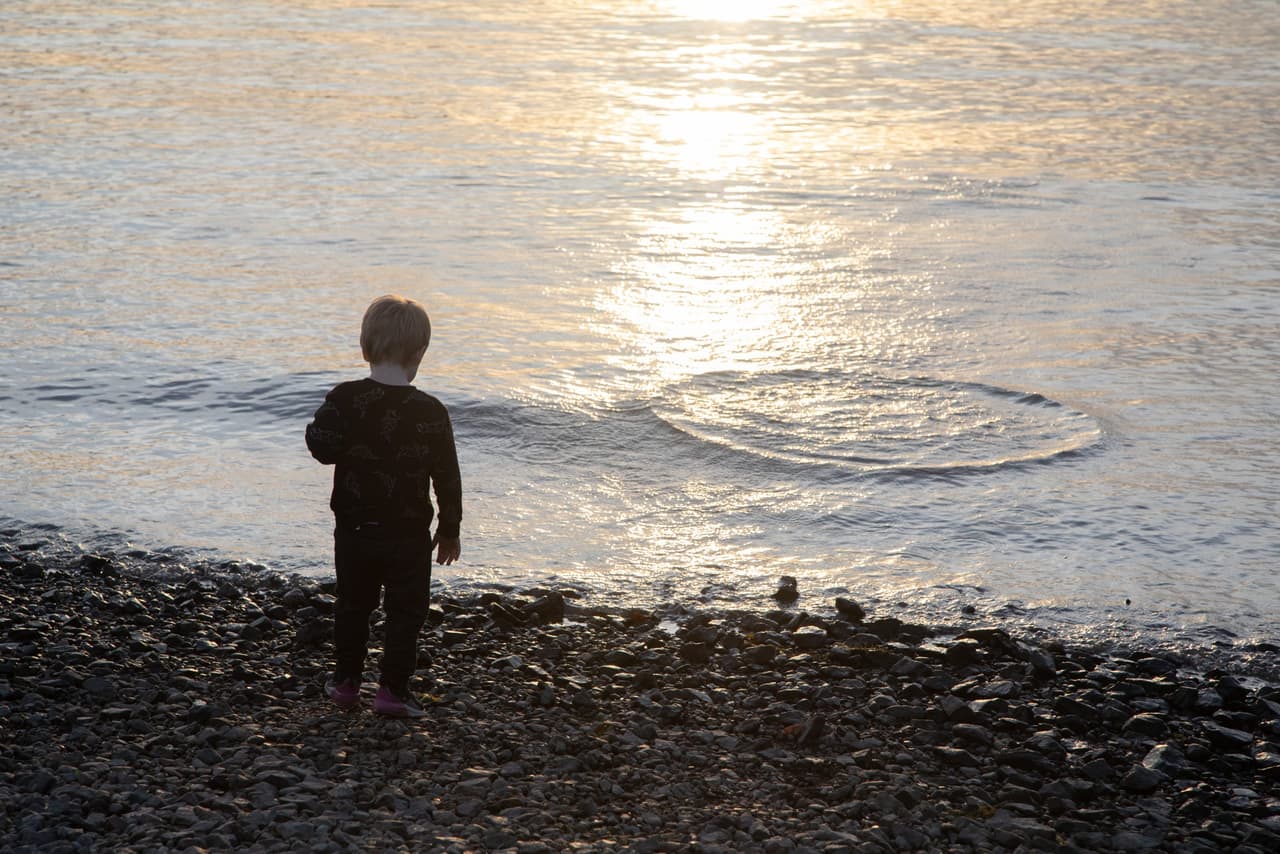 A small child in dark clothes stands alone on a stony river beach, seen from behind, watching a ring of ripples spread across water lit silver by the low sun.