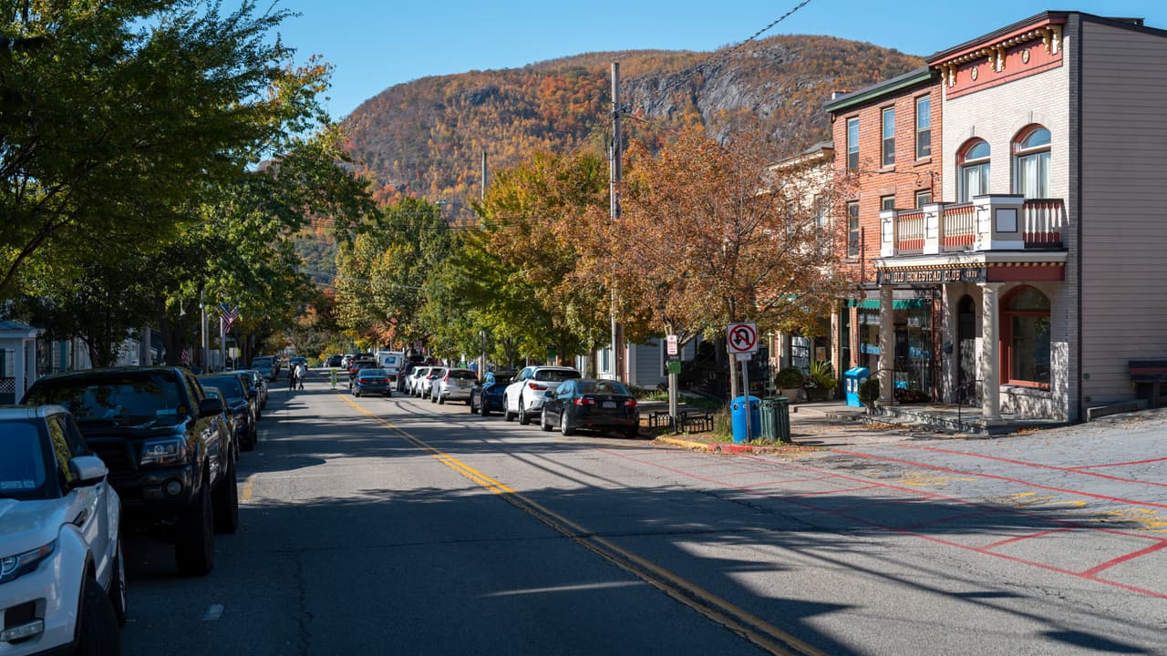 Main Street in the village of Cold Spring on an autumn afternoon: parked cars along both kerbs, two- and three-storey brick and clapboard shopfronts on the right, and the wooded bulk of Storm King closing the end of the street.