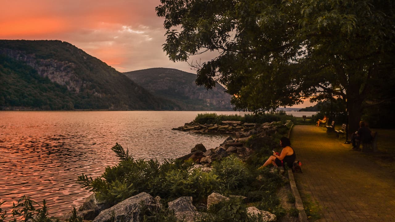 Dusk on the Cold Spring waterfront: a woman sits on the boulders at the water line with her phone, a small dog beside her, under a tree overhanging the brick promenade; a few people rest on benches behind, and the sky above Storm King and the gorge burns pink and orange.