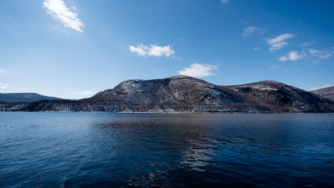 Storm King across the Hudson on a bright winter day: thin snow streaks the bare wooded slopes down to the rail line at the shore, and deep blue rippled water fills the foreground under a clear sky.