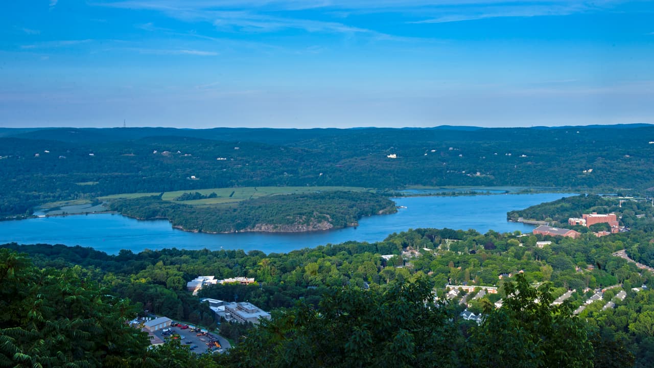 Constitution Island from the US 9W overlook on a clear summer day: the low wooded island reaching into a wide bend of the Hudson, marsh flats behind it, the brick buildings of the northern West Point campus at the right shore, and hazy ridges running to the horizon.