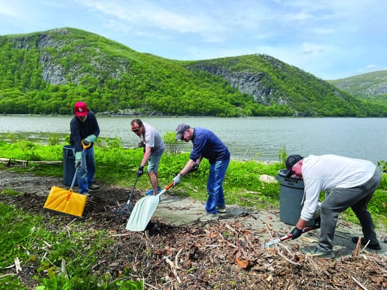 Four volunteers with shovels, rakes and collection bags clearing driftwood and litter from a green riverbank at Dockside Park, with the mountains of the west shore behind them.