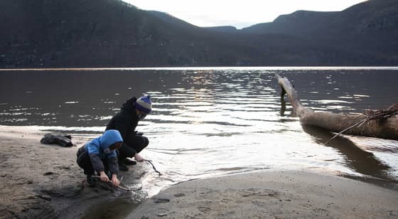 An adult and a child crouch together at the edge of the river on a sand and shingle shore in winter, looking into the shallows, a bleached driftwood log beside them and a dark mountain across the water.