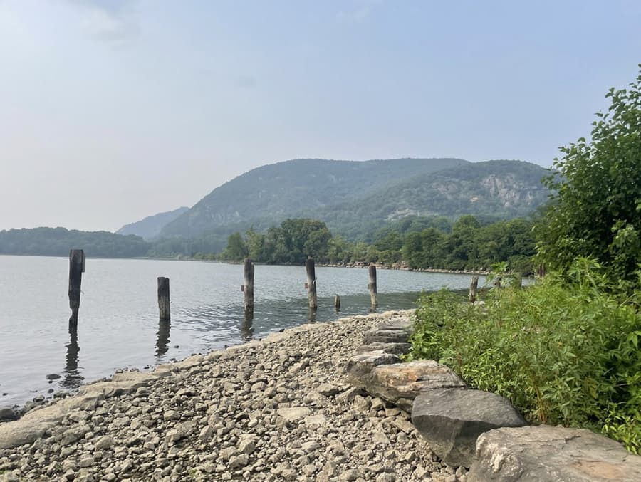 The view north from the shoreline at Dockside Park: a bank of rounded cobbles and boulders running away to the right, a row of weathered timber piles standing in the shallows, open water beyond them, and the wooded mountains upriver in summer haze.