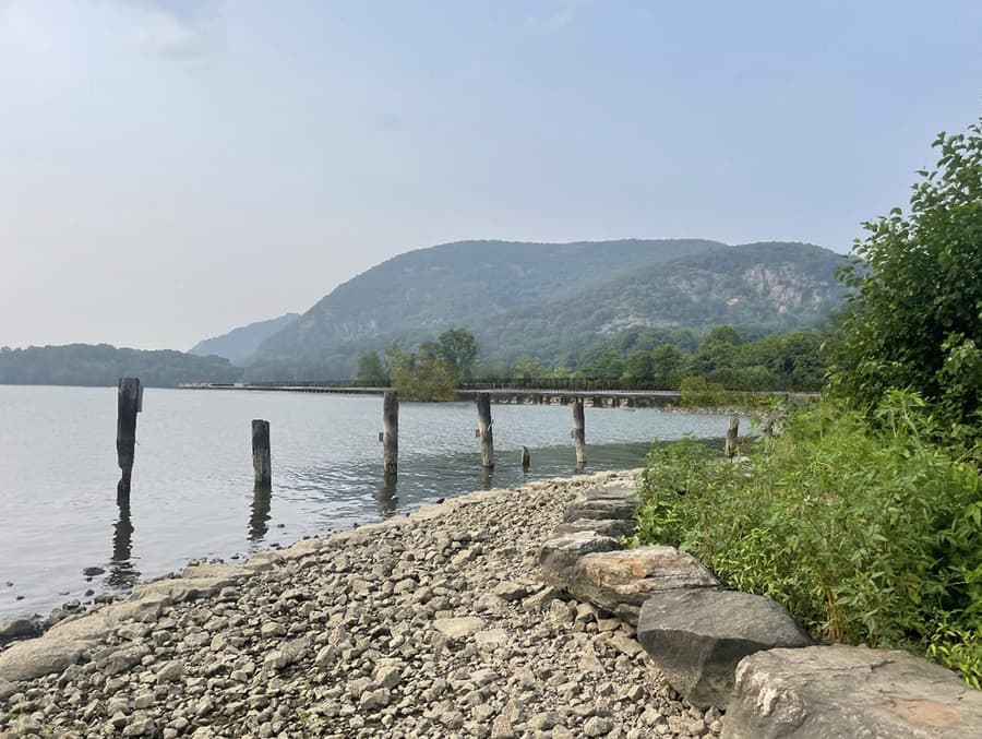 The same view north from Dockside Park with the proposed Fjord Trail built. A long, low horizontal line now crosses the middle distance close to the waterline — the elevated trail deck and its fence running along the causeway — with the cobble bank, the old piles and the mountains otherwise unchanged.