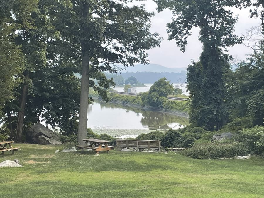 The view south-southwest from higher up at the Little Stony Point stage: mown grass and picnic tables in the foreground under tall trees, a timber rail fence, and the lily-pad inlet with the Metro-North causeway beyond it.