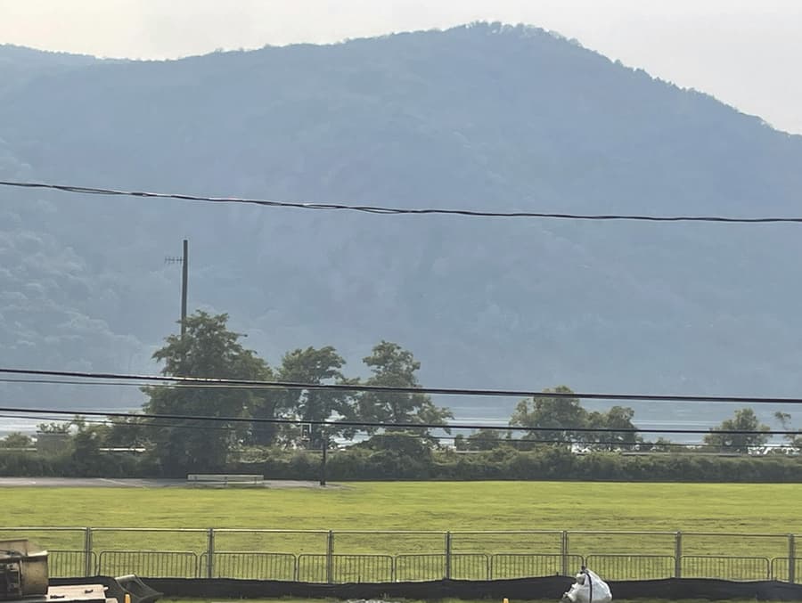 The view west from the residences on lower Fair Street in Cold Spring: grass and scrub in the foreground, a utility pole and a group of small trees at the shoreline, the Hudson beyond, and the wooded mass of Storm King filling the right of the frame.