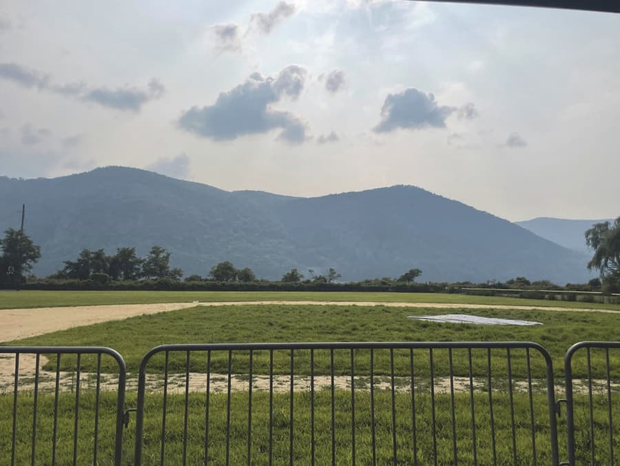 The view west from the playing field at Mayor's Park in Cold Spring: a mown green field behind steel crowd barriers, a line of low trees and the Metro-North tracks across the far edge, and the long ridge of the western Highlands beyond the river under a hazy summer sky.