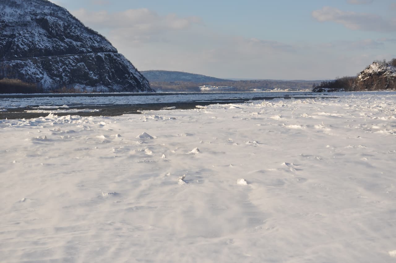 The Hudson at Cold Spring frozen over, broken white ice filling the whole channel from bank to bank, with the snow-streaked flank of Storm King rising into flat grey winter light beyond.
