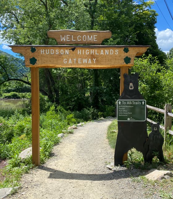 A timber gateway arch over a gravel path reading "Welcome — Hudson Highlands Gateway", with a carved black bear beside the right-hand post and a green trail information panel behind it. Summer woodland either side.