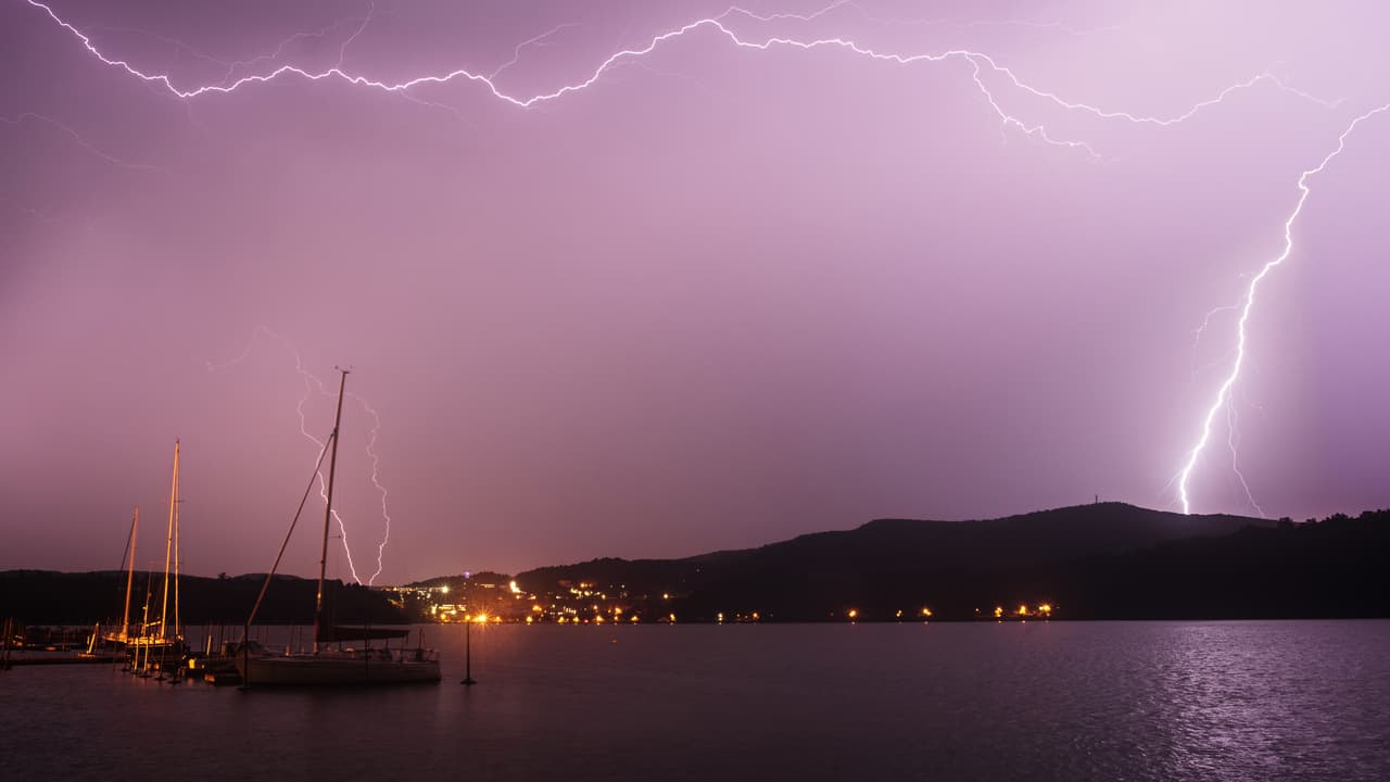 A night thunderstorm over the Hudson at Cold Spring: two long lightning bolts fork across a violet sky and one strikes behind the dark ridge on the far shore, while sailboats sit at their moorings and the lights of the west bank reflect in the water.