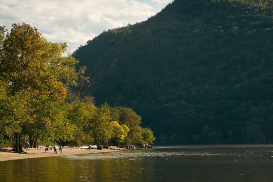 The gravel beach at Little Stony Point, with a handful of people at the water line and the steep wooded shoulder of the Highlands rising straight out of the river behind, trees just turning.