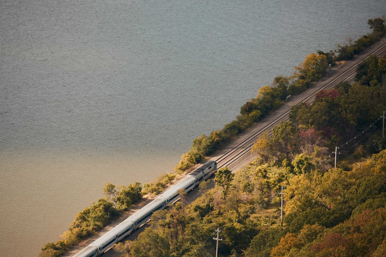 Looking down from above on a Metro-North train running north along the Hudson shoreline, the track pinched between the water on one side and a steep autumn hillside on the other.