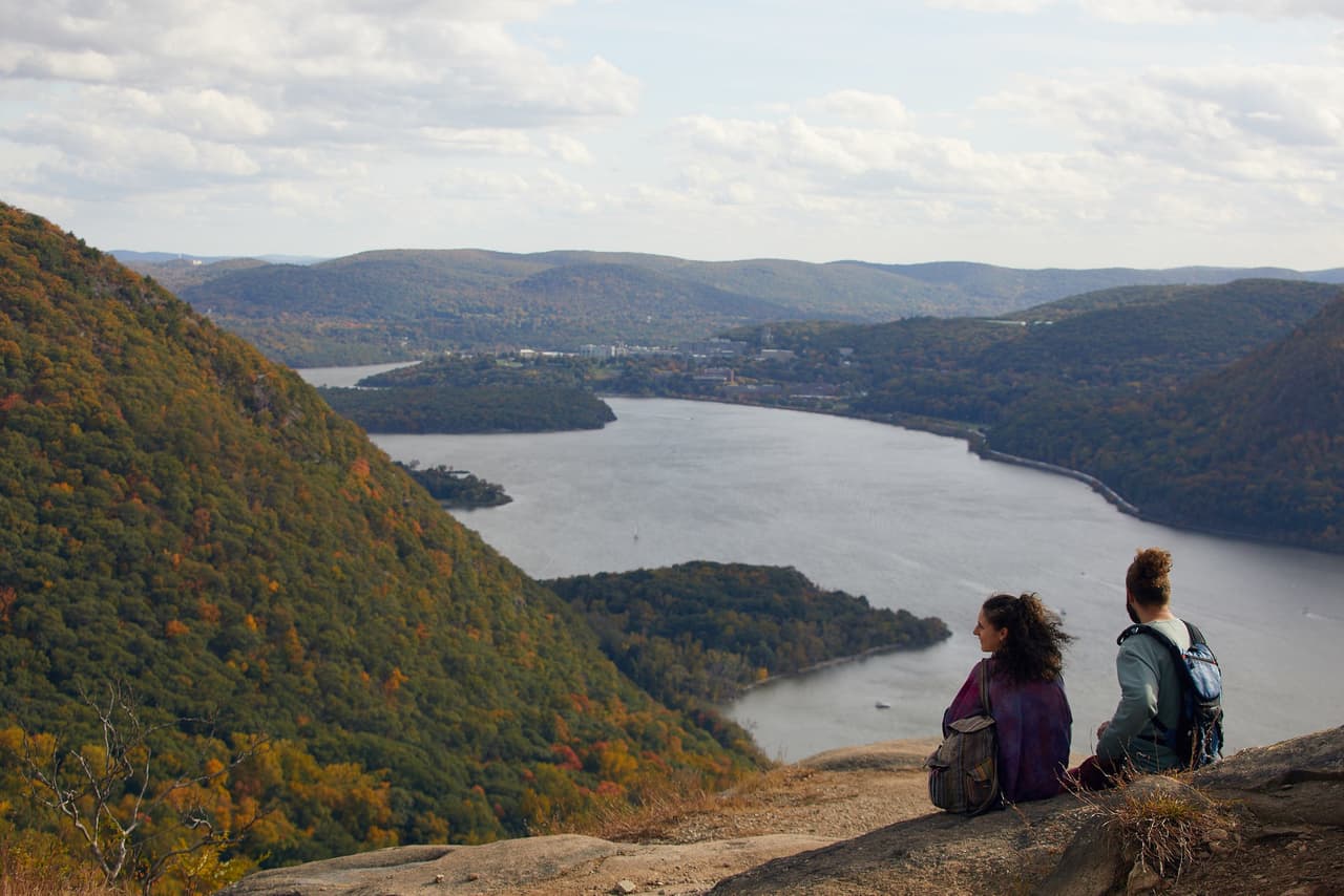 Two people sit close together on a bare rock ledge high above the Hudson, backs to the camera, looking down the river as it runs between wooded ridges in autumn colour.