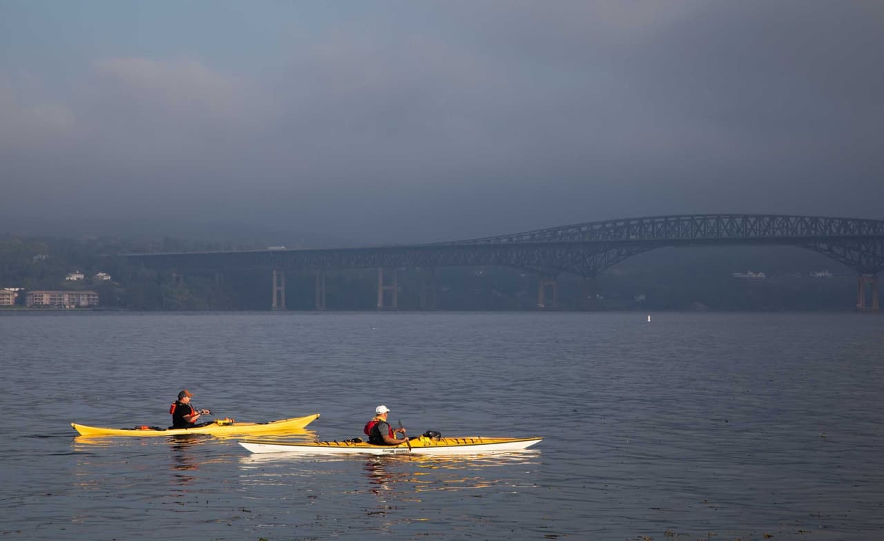 Two yellow sea kayaks crossing flat grey water on an overcast day, small against the long low span of the Newburgh–Beacon Bridge in the haze behind them.