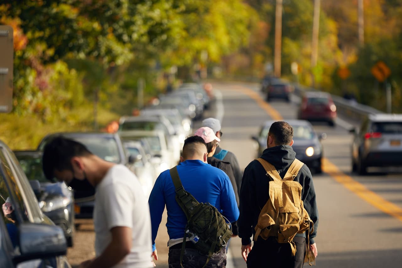 Four people with backpacks walking in the traffic lane of Route 9D, threading between a line of parked cars on one side and oncoming vehicles on the other.