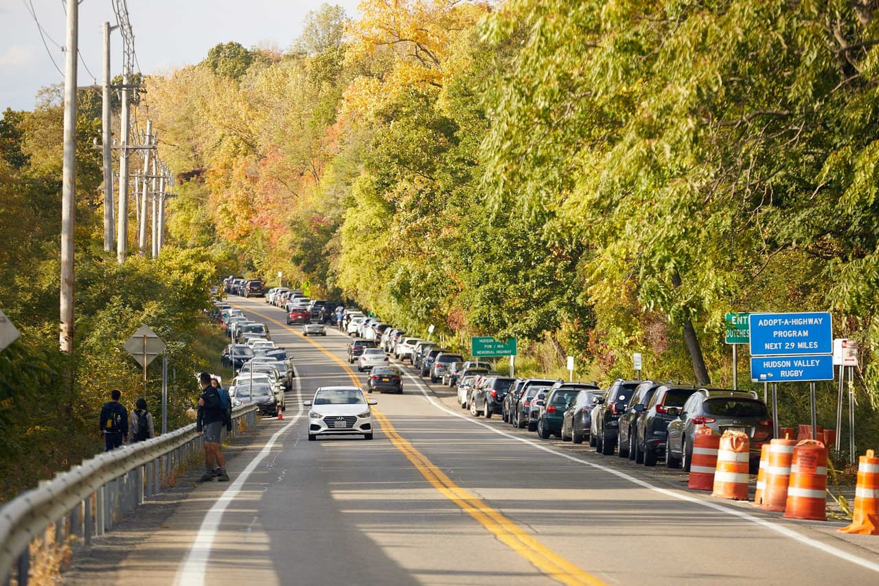 Route 9D on a peak autumn weekend: cars parked nose to tail along both shoulders for as far as the road runs, orange traffic barrels narrowing the carriageway, and people walking in the roadway between the parked cars and moving traffic.