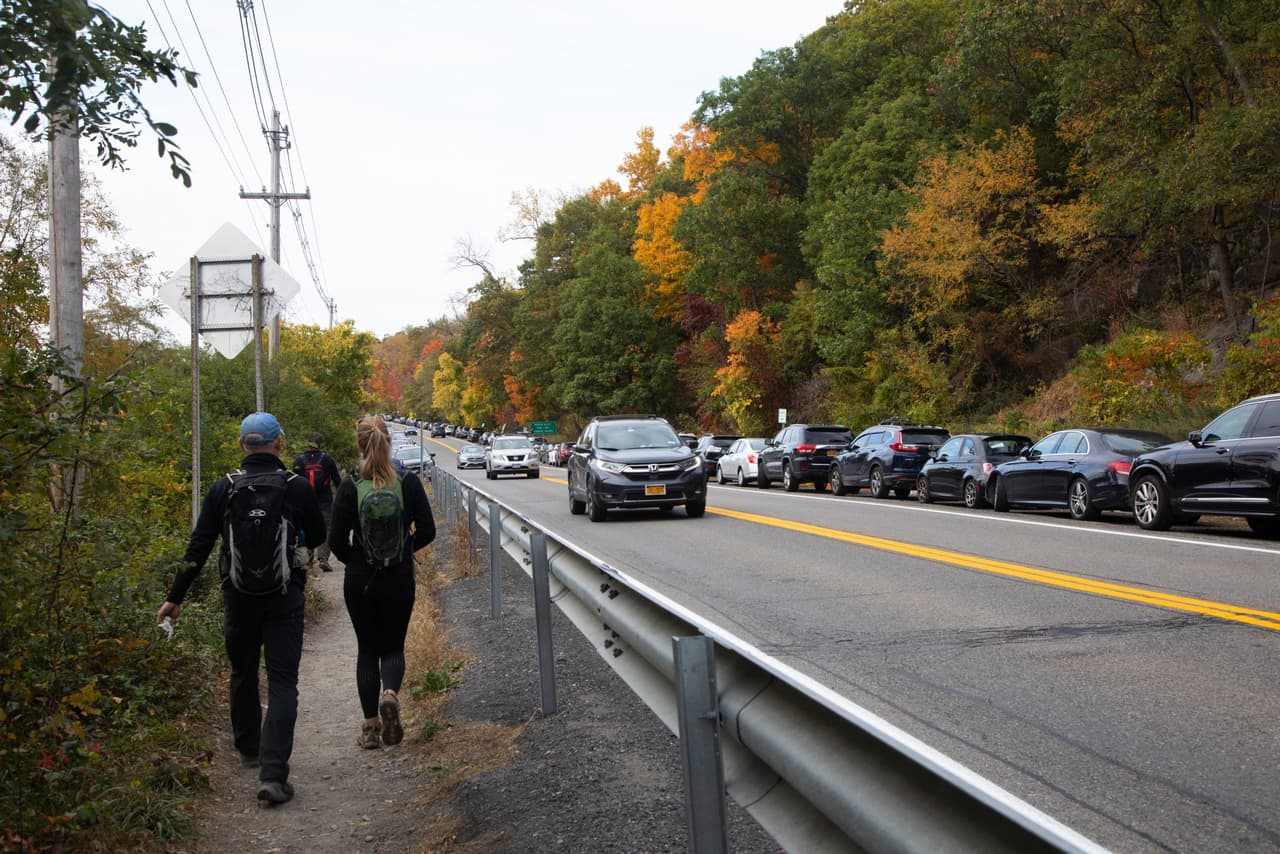 Two hikers with daypacks walk single file along a strip of dirt outside the guardrail on Route 9D, level with a solid line of parked cars and a lane of moving traffic. Autumn colour on the slope above the road.