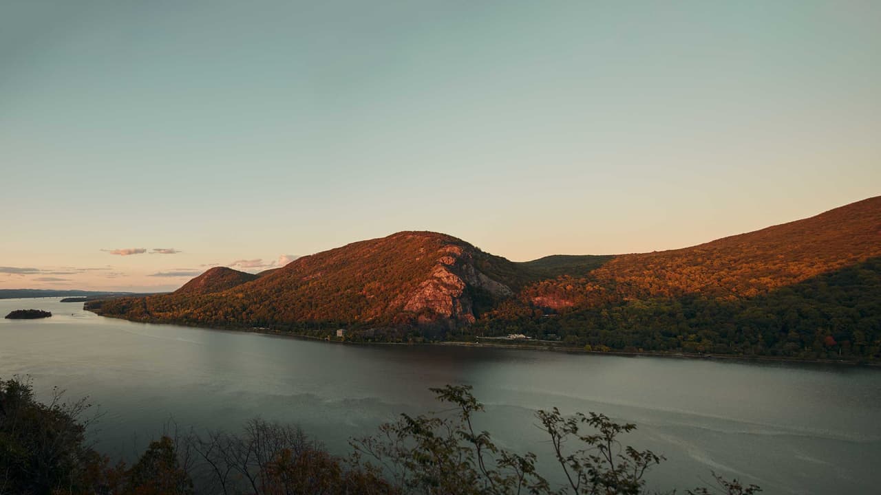 The fjord from high ground at sunset: the last light turns the bare rock face of Breakneck Ridge deep orange above autumn woodland, the river lies flat and pale below, and Pollepel Island and the Newburgh-Beacon Bridge fade into the haze upriver.