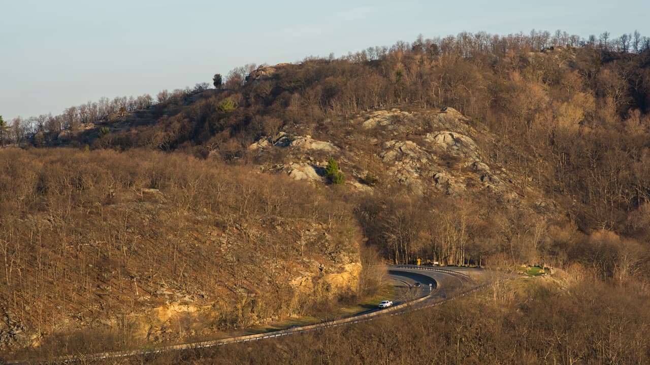 US Route 9W curving around the shoulder of Storm King State Park in late-day light: a white pickup on the two-lane road below a rock cut, leafless early-spring woods covering the slope, and bare grey outcrops breaking through the ridge above.